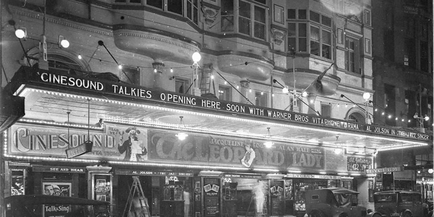 A cinema front at night in Sydney in 1928 with signs announcing that talkies are coming soon